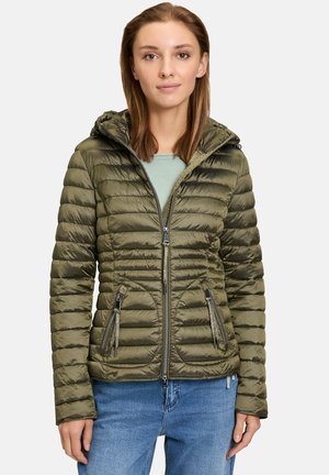 Veste matelassée vert olive avec capuche, fermeture éclair à l'avant, deux poches latérales zippées et texture en matelassage. Portée avec un haut bleu clair et un jean.