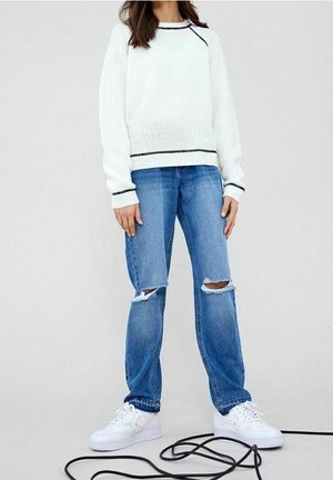 Henkilö, joka pukeutunut valkoiseen neulepuseroon mustilla reunoilla, revittyihin sinisiin farkkuihin ja valkoisiin lenkkareihin, seisoo mustan kaapelin vieressä vaalealla taustalla.