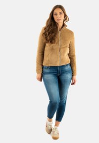 Giacca zip-up marrone, testurizzata, con colletto alto, jeans aderenti blu e sneakers beige con dettagli rossi. Modello in piedi su uno sfondo neutro.
