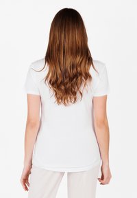 T-shirt bianco in cotone a maniche corte con orlo arrotondato, indossato. Lunghi capelli castano ondulati visibili, in piedi contro uno sfondo bianco uniforme.
