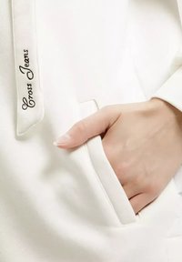 Biała bluza z kapturem z boczną kieszenią, z haftem "Cross Jeans" w ciemnym kolorze po lewej stronie, blisko otworu.