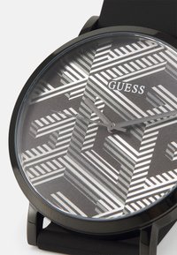 Schwarze Uhr mit einem runden Zifferblatt, das ein geometrisches Streifenmuster in Grau und Weiß zeigt, sowie einer dezenten Zweizeigerbewegung. Der Markenname "GUESS" ist angezeigt.