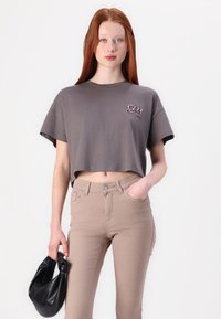 Gray cropped t-shirt featuring a colorful logo on the chest, paired with light beige fitted jeans. Model holds a black handbag.