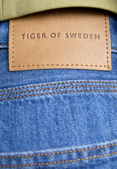 Tela de denim azul con costuras visibles. Tiene una etiqueta de cuero marrón cosida, con el texto "TIGER OF SWEDEN" en relieve. Un tejido verde es parcialmente visible en la parte superior.