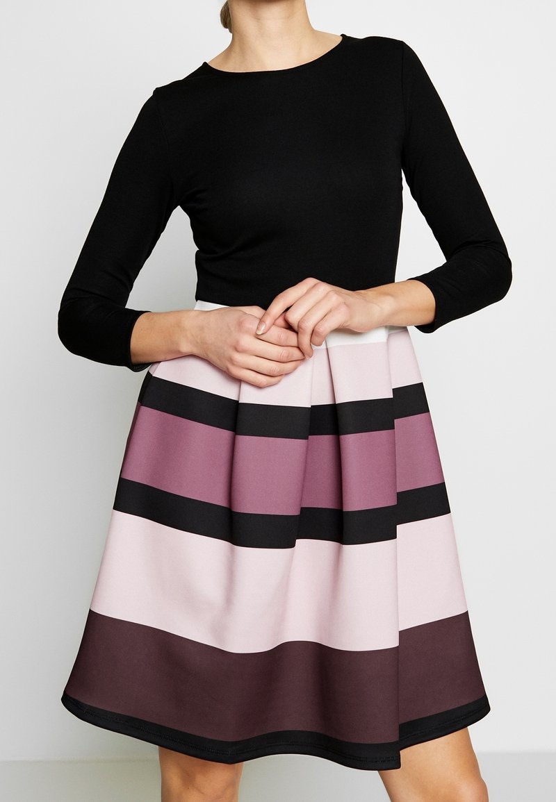 Femme portant un haut noir à manches longues avec une jupe plissée aux rayures horizontales dans des tons de violet, rose, noir et marron.