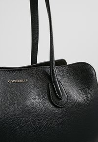 Sac à main en cuir texturé noir avec deux anses et petit logo doré de la marque Coccinelle en haut à gauche.