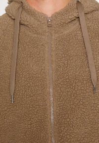 Felpa con cappuccio marrone in pile, con chiusura a zip, esterno testurizzato, cordini regolabili e dettagli metallici sulla zip.