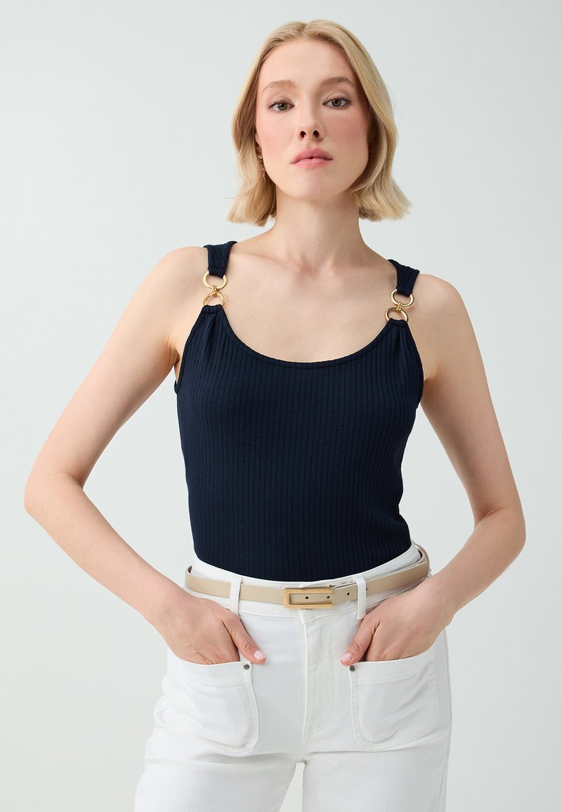 Femme aux cheveux blonds courts portant un haut sans manches bleu marine avec des anneaux dorés sur les bretelles et un pantalon blanc avec une ceinture beige, les mains dans les poches.