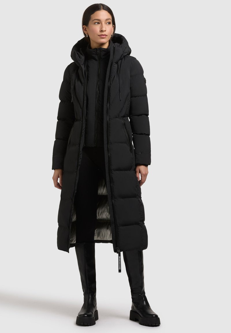Abrigo largo acolchado negro con capucha, con cierre de cremallera, bolsillos laterales y un forro suave. Lleva puesto sobre una camiseta negra y se combina con botas altas hasta la rodilla.