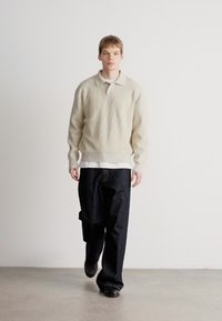 Pulover ribbed de culoare bej deschis cu guler, purtat peste o cămașă albă. Blugi largi de culoare bleu închis, pantofi negri, fundal minimalistic.