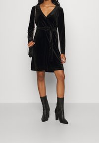 Svart sammetsomslagsklänning med långa ärmar, bälte i midjan, V-ringning och knälängd, kombinerad med svarta klackstövlar. 