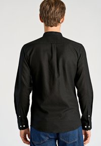 Czarna koszula z długim rękawem o luźnym kroju, z mankietami na guziki i szwem na plecach, wykonana z teksturowanej tkaniny, zestawiona z niebieskimi dżinsami.