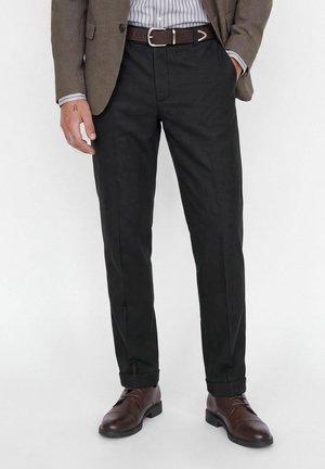 Homme portant un pantalon de costume noir, une ceinture marron foncé, des chaussures en cuir marron, une chemise rayée et un blazer marron avec une main dans la poche.