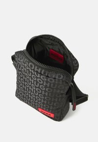 Sac bandoulière noir compact avec impression répétée du logo "HUGO", étiquette rouge avec logo, et fermeture éclair ouverte montrant un intérieur noir avec un patch logo rouge.