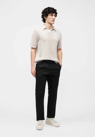 Homme aux cheveux bouclés portant un polo en maille beige à manches courtes, pantalon noir, baskets blanches, debout les mains dans les poches devant un fond uni.