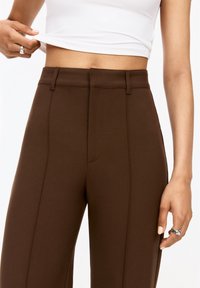 Pantalon marron sur mesure en tissu lisse, doté d'une taille haute, de plis à l'avant et d'une finition nette. Des mains ajustent un haut blanc.