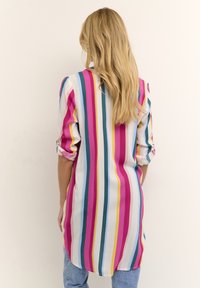 Chemise à manches longues rayée en rose, bleu, jaune et blanc. Tissu lisse avec des manches retroussées et un ourlet arrondi, associée à un jean en denim.