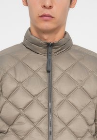 Gepolsterte, hellgraue Jacke mit hohem Kragen, Frontreißverschluss und einer strukturierten Rautenoptik. Weiches, synthetisches Material und minimalistische Hardware-Details.