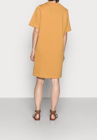 Persona de pie de espaldas, vestida con un vestido mostaza de longitud a la rodilla y sandalias marrones con tiras al tobillo, sobre un fondo liso.