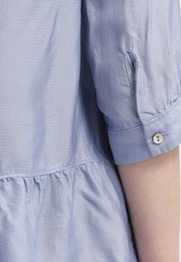 Ljusblå blus med texturerad tyg, uppvikta ärmar och knäppning framtill. Inkluderar rynkdetalj vid midjan för extra struktur.