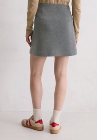 Grauer ausgestellter Rock aus gewebtem Stoff, kombiniert mit beigefarbenem Langarmoberteil, cremefarbenen Socken und beigen Schuhen mit roten Akzenten. Sichtbare gepunktete Textur.