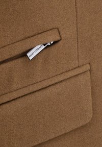 Brązowa wełniana tkanina o gładkiej fakturze z kieszenią i srebrnym zamkiem błyskawicznym oraz metką marki wszytą w szew dla szczegółu.