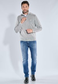 Grau Fleece-Pullover mit Reißverschluss-Kragen, versehen mit einem Logo auf der Brust. Getragen mit blauen Jeans und schwarzen Schnürstiefeln, vor einem hellen Hintergrund.