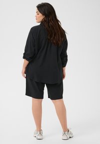 Camisa negra de botones con mangas enrolladas combinada con pantalones cortos negros. El conjunto tiene un ajuste relajado y se complementa con zapatillas de color claro.