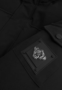 Zwarte gevoerde jas met een gestructureerde stof, een glanzend logopatch en versterkte stiksels. Het heeft een strakke, moderne uitstraling met opvallende hardware-accenten.