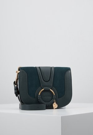 Ciemnozielona torebka na ramię z skóry i zamszu z złotym pierścieniem i plecionym detalem, regulowanym paskiem oraz małym złotym breloczkiem na białym tle.