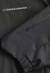 Veste coupe-vent noire avec une texture lisse, des poignets élastiques et une poche zippée. Le logo de la marque est embossé sur le devant.