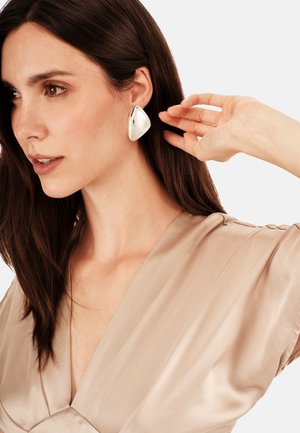 Stříbrné prohlášení náušnice s lesklým, slzovitým tvarem. Modelka má na sobě béžový saténový top, který zvýrazňuje lesk a design doplňku.