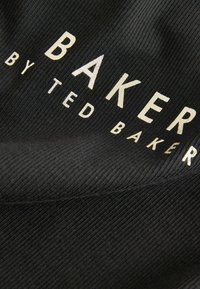 Zwarte ribstof met de tekst "BAKER" in grote gouden letters en "by TED BAKER" in een kleinere lettergrootte, met een gladde textuur en een lichte glans.