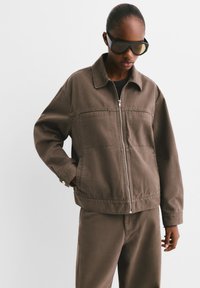 Veste en coton marron avec fermeture éclair à l'avant, poches latérales et col. Le modèle porte des lunettes de soleil ; l'ensemble comprend un pantalon assorti.