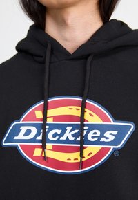 Osoba v černé mikině s kapucí a barevným logem Dickies v červené, žluté a modré barvě na hrudi, ukazující část krku a vlasů.