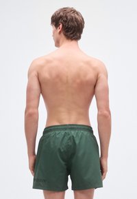 Zaļas peldēšanas šorti ar elastīgu vidukli un logotipu kreisajā pusē. Audums ir gluds un spīdīgs.