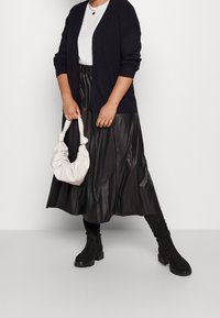 Cárdigan negro de punto sobre una camiseta blanca, combinado con una falda midi de cuero sintético negra con pliegues. Lleva un bolso color crema y botas negras.