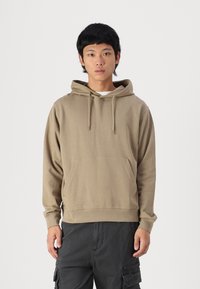 Beige hoodie i mjukt tyg, med en framficka, justerbar dragsko och ribbade ärmslut. Bärs med mörka cargobyxor.