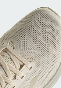 Lichtbeige atletische schoen met een gebreide bovenkant met textuur, voorzien van kleine ventilatiegaatjes en een plat vetersluitingssysteem.