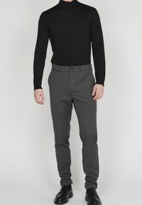 Man som bär svart polotröja, gråa smal passformbyxor och svarta läderloafers, stående mot en plain ljus bakgrund.