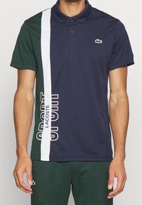 Man som bär en marinblå och grön polo med vita vertikala ränder och texten "Lacoste Sport", som är parat med mörkgröna shorts med en liten logotyp.