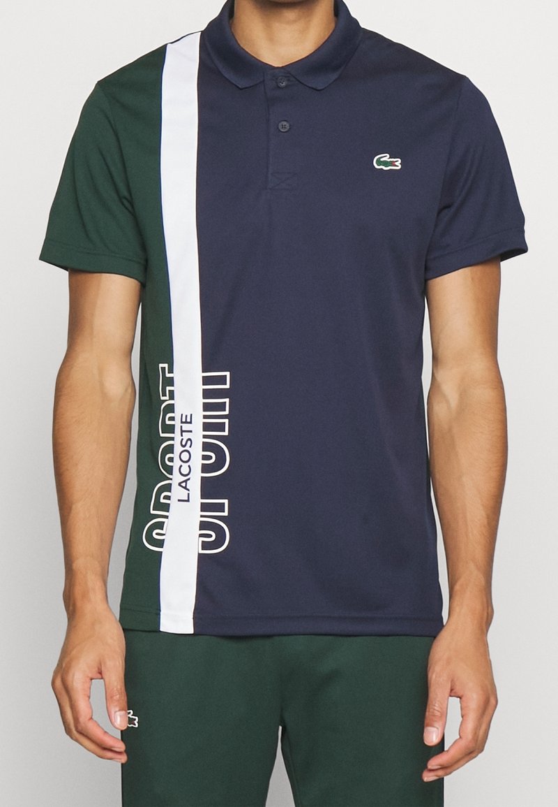 Man som bär en marinblå och grön polo med vita vertikala ränder och texten "Lacoste Sport", som är parat med mörkgröna shorts med en liten logotyp.