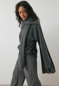 Vrouw met lang, donker krullend haar, gekleed in een bijpassende donkergrijze trui en sjaal, en losse, getextuurde grijze broek, staand tegen een rustige muur.