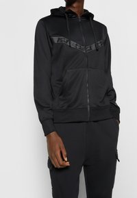 Sudadera con cremallera negra de acabado suave, con un panel contrastante en el pecho, bolsillo tipo canguro y capucha con cordón.