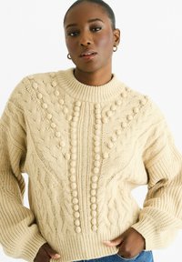 Beige cable-knit sweater with a high ribbed neckline, textured patterns, and raised bobble accents down the center. Relaxed fit design.
