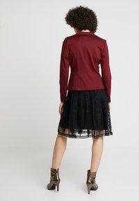 Burgundy-fitattu takki, jossa on lovimallinen kaulus, yhdistettynä mustaan pitsihameeseen, jossa on kerroksellisia kuvioita, sekä ruskeisiin nilkkureihin.