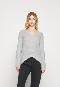 Hellgrauer, strukturierter Strickpullover mit V-Ausschnitt, langen Ärmeln und asymmetrischem Saum. Kombiniert mit schwarzen Jeans.