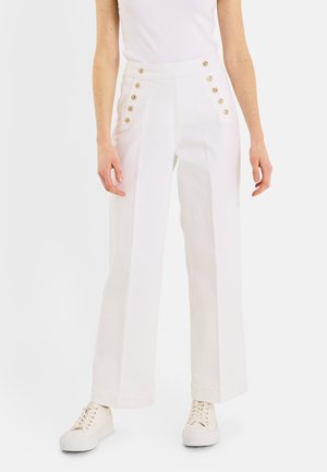 Femme portant un pantalon blanc taille haute à jambes larges avec deux rangées de boutons dorés, des baskets blanches et un top blanc sans manches.