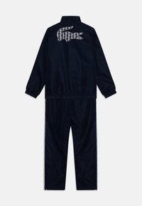 Tamsiai mėlynas sportinis kostiumas, pagamintas iš lygaus audinio, su aukštu apykakle, elastingais rankogaliais ir baltais akcento juostelėmis šonuose. Lacoste logotipas ant nugaros.