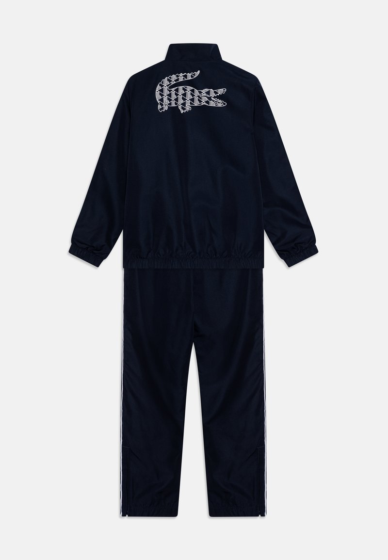 Dunkelblauer Trainingsanzug aus glatt aufgehäuftem Stoff, mit hohem Kragen, elastischen Bündchen und weißen Akzentstreifen an den Seiten. Lacoste-Logo auf dem Rücken.